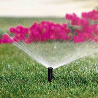 An image of a small sprinkler watering green grass backdropped by pink flowers.